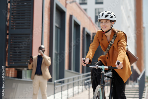 Fototapeta Naklejka Na Ścianę i Meble -  Young cheerful female solopreneur in safety helmet and casualwear sitting on bicycle and riding home from work in urban environment
