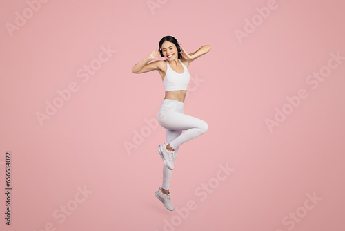 Excited woman in sportswear and wireless headphones jumping over pink background, full length shot, free space