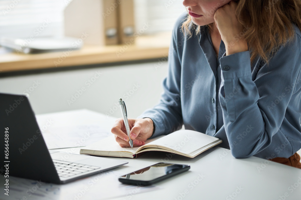Cropped shot of young businesswoman or student in blue shirt holding pen over open notebook while thinking of new working ideas in office Cropped shot of young businesswoman or student in blue shirt holding pen over open notebook while thinking of new working ideas in office