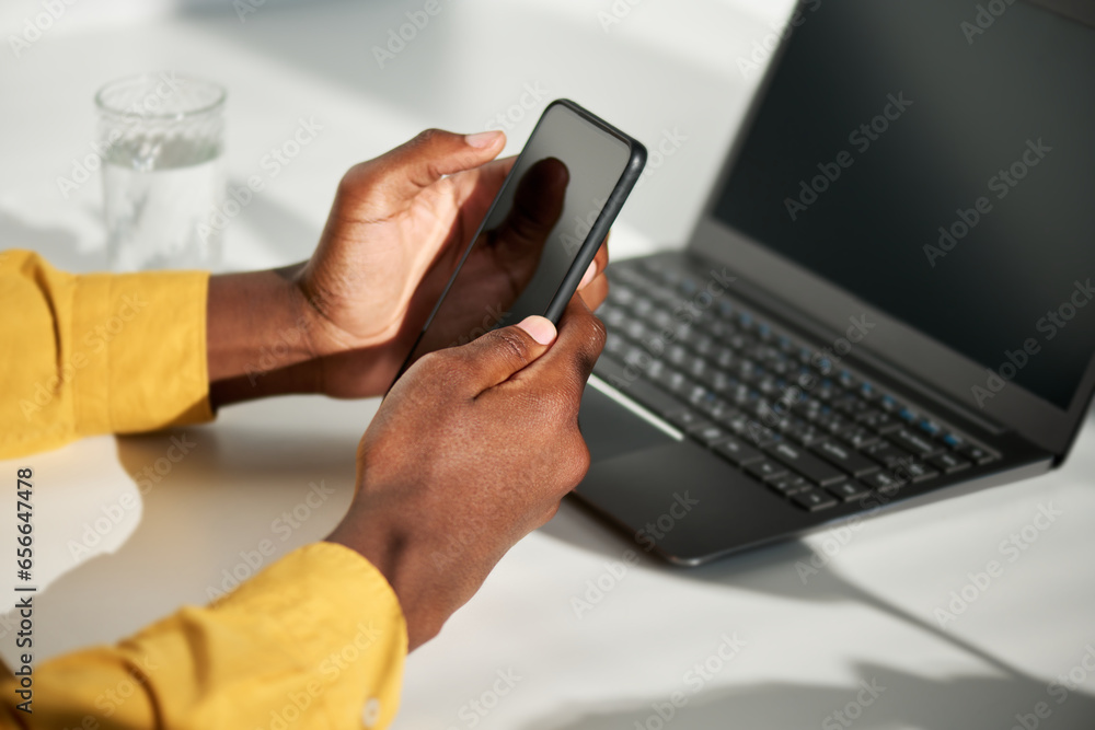 Focus on hands of black businessman with mobile phone texting or scrolling through data while sitting by workplace in front of laptop Focus on hands of black businessman with mobile phone texting or scrolling through data while sitting by workplace in front of laptop