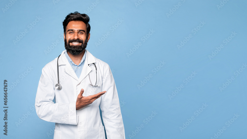 Cheerful indian male doctor therapist in white coat showing free space isolated on blue background, panorama, banner Cheerful indian male doctor therapist in white coat showing free space isolated on blue background, panorama, banner