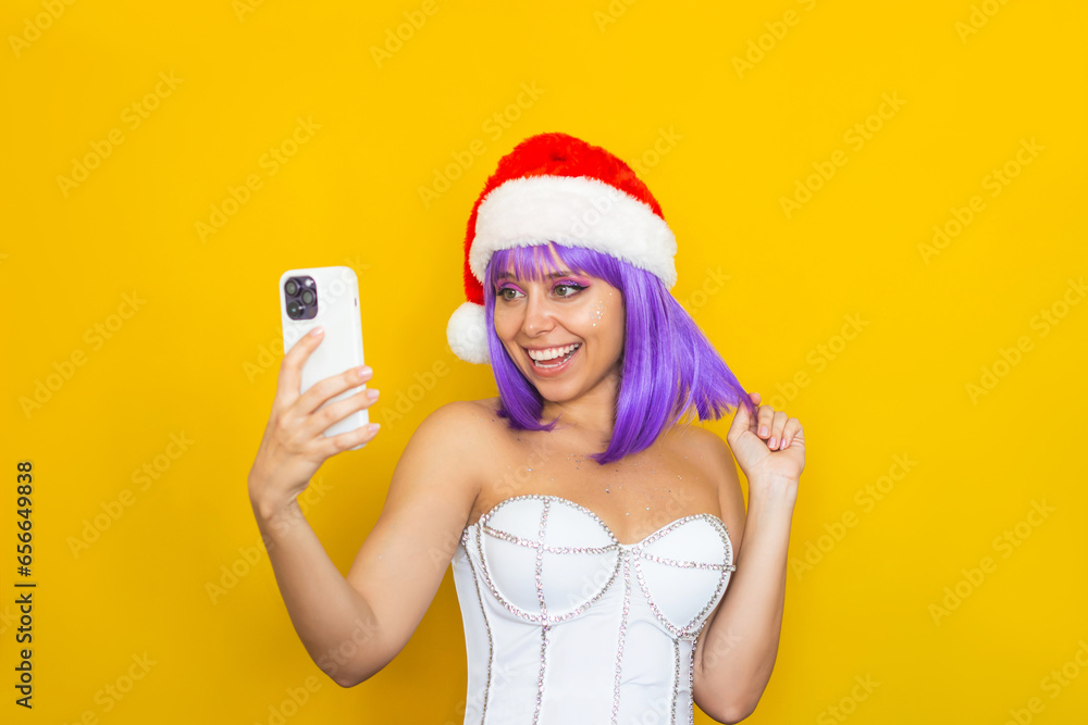 A young happy smiling caucasian woman with purple hair in white festive corset, red Santa Claus hat takeing a selfie with her mobile phone isolated on color yellow background. Christmas and New Year A young happy smiling caucasian woman with purple hair in white festive corset, red Santa Claus hat takeing a selfie with her mobile phone isolated on color yellow background. Christmas and New Year