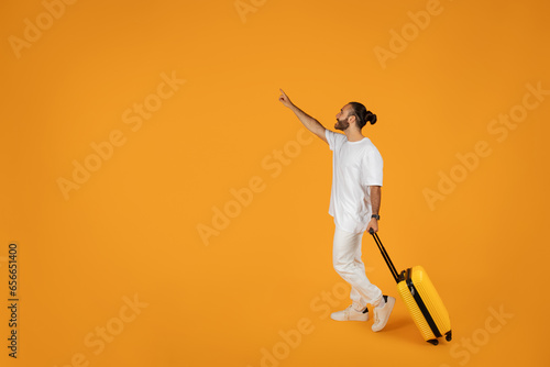 Smiling shocked adult european guy in white t-shirt with suitcase points finger at empty space photo