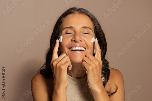 Happy caucasian elderly woman apply cream on face, enjoy routine and spa procedures photo