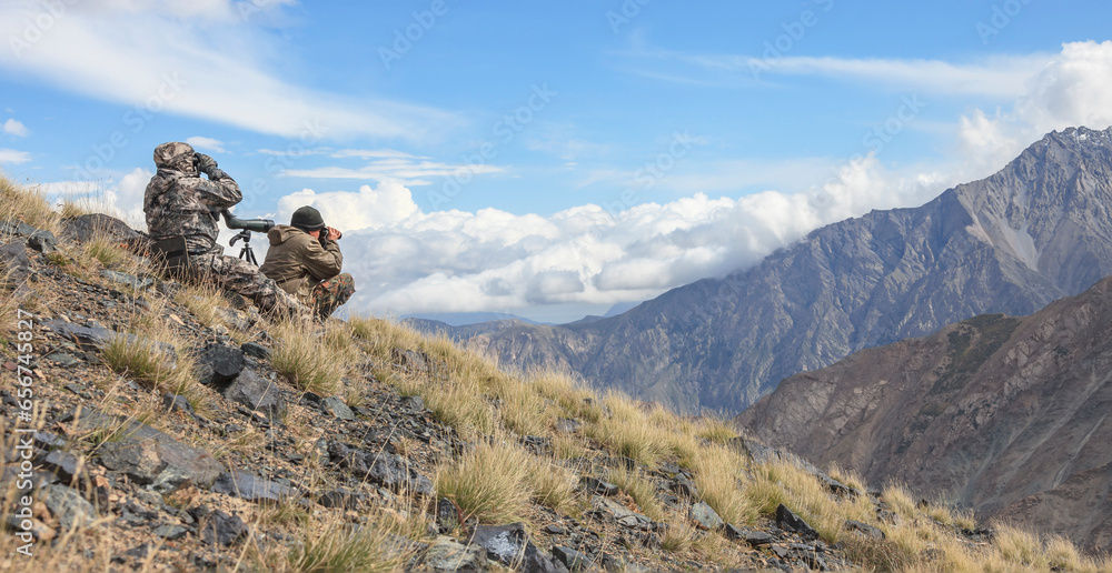 Couple of men in camouflage with binoculars and telescope are observing in the high mountains.