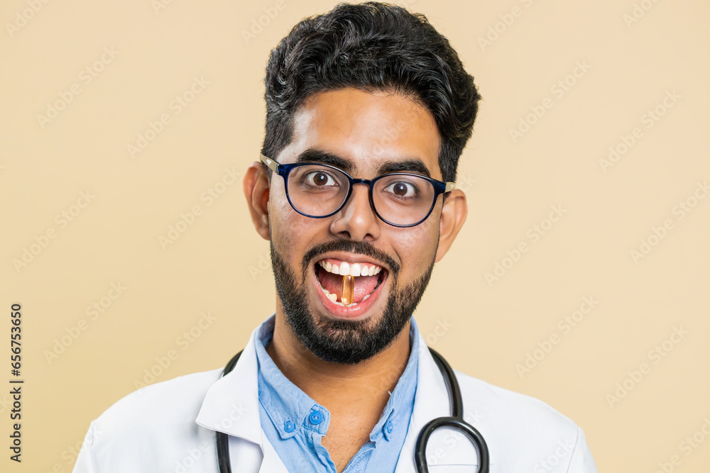 Smiling Indian doctor man puts of tablet pill vitamin D or omega-3 yellow capsule into mouth, recommends drugs immunization cure treatment. Health care. Apothecary pharmacist on beige background Smiling Indian doctor man puts of tablet pill vitamin D or omega-3 yellow capsule into mouth, recommends drugs immunization cure treatment. Health care. Apothecary pharmacist on beige background
