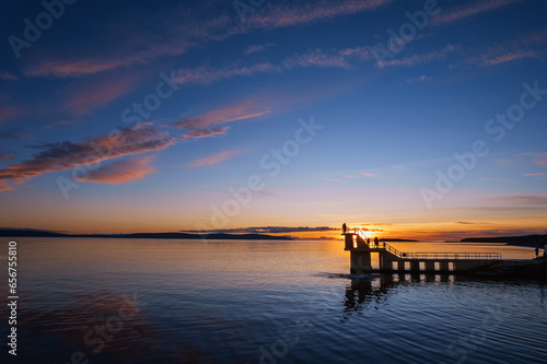 Silhouette of Black rock diving board with people looking at stunning nature sunset scene. Salthill beach, Galway town, Ireland. Rich blue and orange color. Famous city landmark and area. Aerial view.