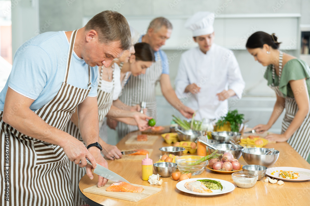 Focused man in apron standing at table with groceries and utensils during group cooking lesson, absorbed in cooking process, cutting fish following chef instructions..
