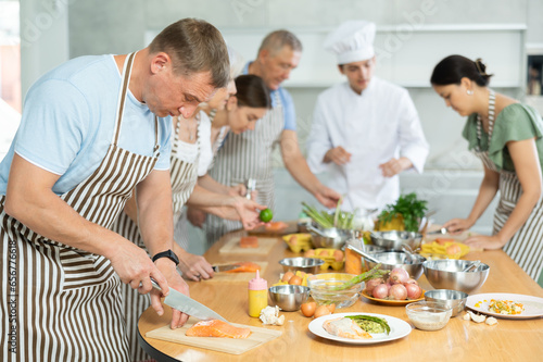 Focused man in apron standing at table with groceries and utensils during group cooking lesson  absorbed in cooking process  cutting fish following chef instructions..