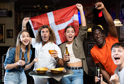 Group of Denmark football team fans spending time in bar, drinking bear and having fun. People with state flag in pub.