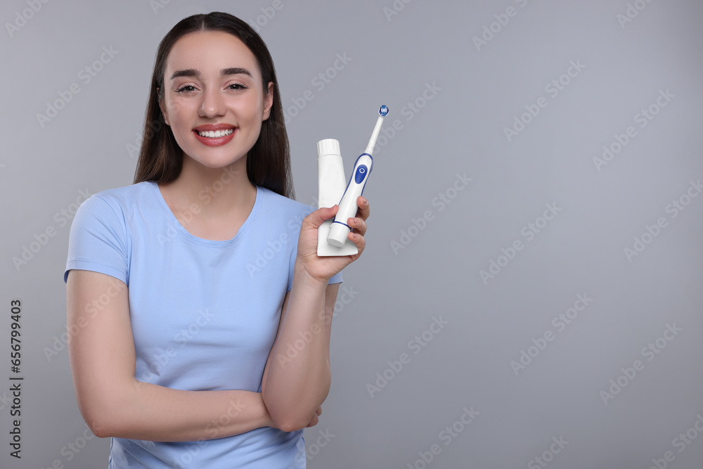 Happy young woman holding electric toothbrush and tube of toothpaste on light grey background, space for text Happy young woman holding electric toothbrush and tube of toothpaste on light grey background, space for text