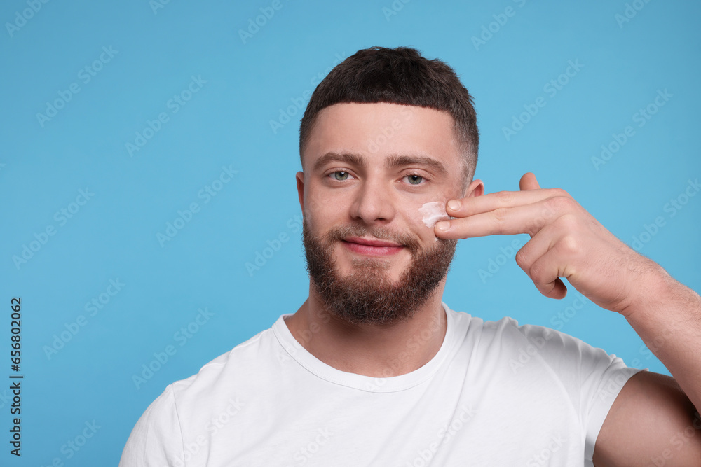 Handsome man applying cream onto his face on light blue background Handsome man applying cream onto his face on light blue background