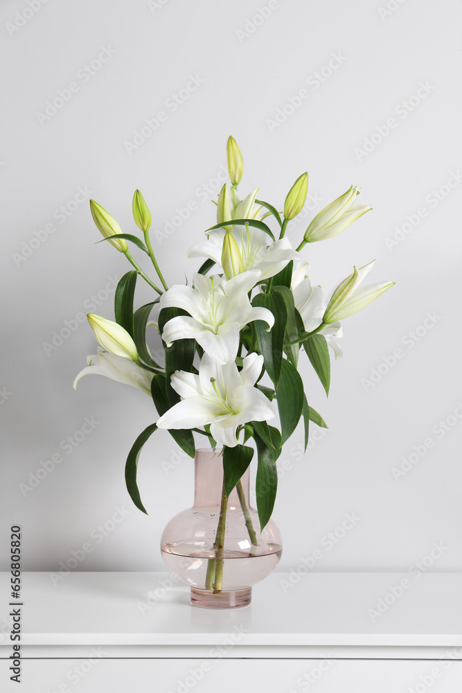 Beautiful bouquet of lily flowers in glass vase on table near white wall