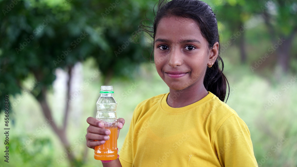 Happy Cute Indian girl drinking juice in the park, holding plastic bottle and showing thumb up. outdoor shot Happy Cute Indian girl drinking juice in the park, holding plastic bottle and showing thumb up. outdoor shot