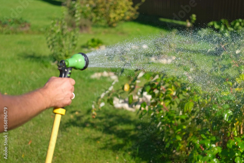 Gardener with a watering hose. Selective focus on a drops. Person spraying green grass lawn with sprayer. Irrigation with water, sunny day. Garden sprinkler. Landscaping. Gardening, waters plants care