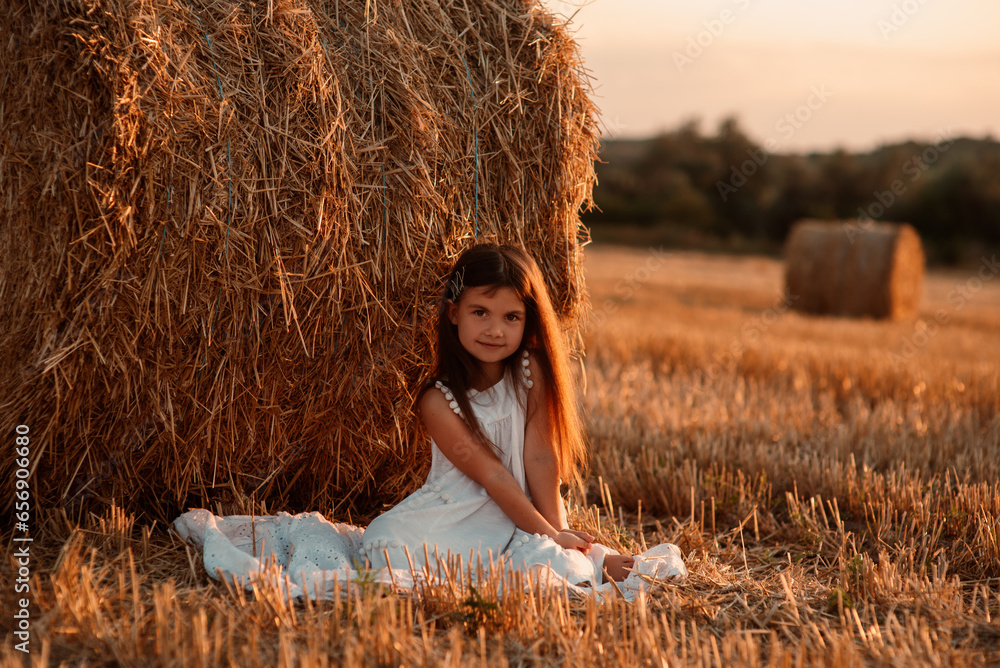 little girl in white dress spending time in a field of cut wheat among the bales at sunset little girl in white dress spending time in a field of cut wheat among the bales at sunset