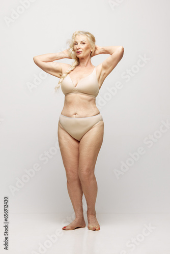 Full-length of elegant, beautiful, senior woman with healthy, natural body posing in underwear against grey studio background. Concept of age, natural beauty. body and skin care, healthy lifestyle