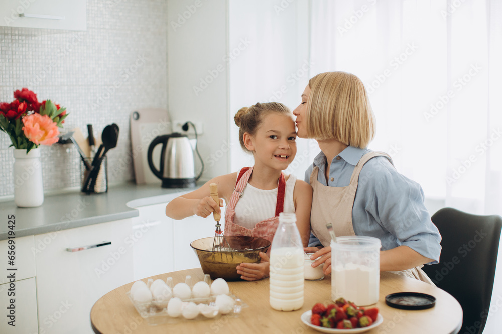 Mother and daughter are preparing pancake batter at the table in a modern kitchen. The girl holds a whisk, stirs eggs in a bowl, prepares dough with her mother. Mother and daughter are preparing pancake batter at the table in a modern kitchen. The girl holds a whisk, stirs eggs in a bowl, prepares dough with her mother.