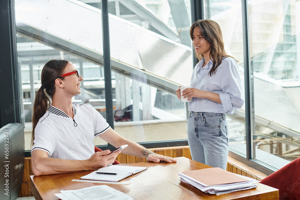 Fototapeta premium cheerful young associates talking and smiling sincerely holding tea cup and phone, coworking