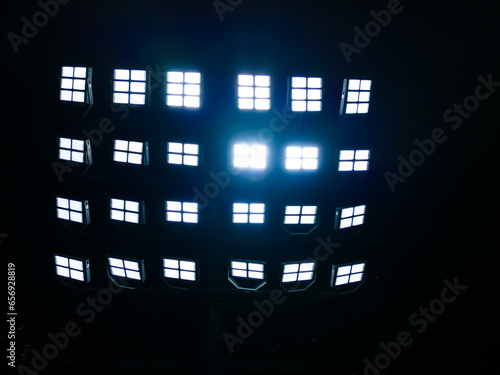 A close-up of stadium floodlights, illuminating the field. Powerful beams of light pierce the darkness, creating a sense of energy and excitement.