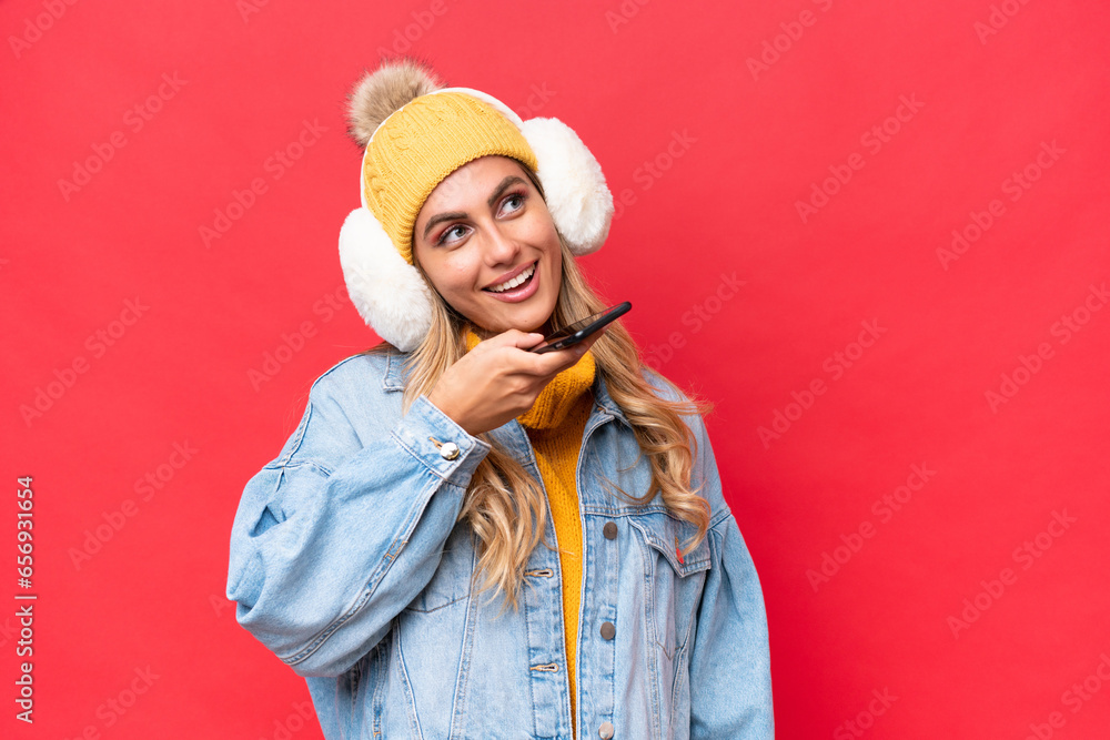 Young pretty Uruguayan woman wearing winter muffs isolated on red background background keeping a conversation with the mobile phone