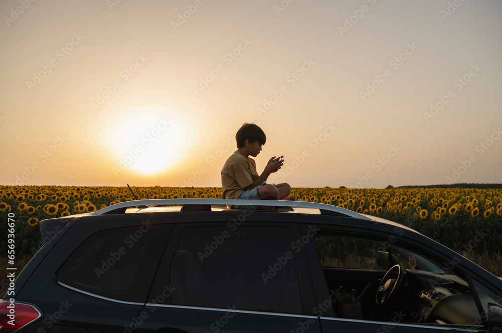 Boy using smart phone sitting on top of car at sunset Boy using smart phone sitting on top of car at sunset