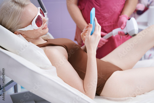 Woman using smart phone getting laser epilation treatment at clinic photo