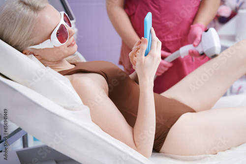 Woman using smart phone getting laser epilation treatment at clinic