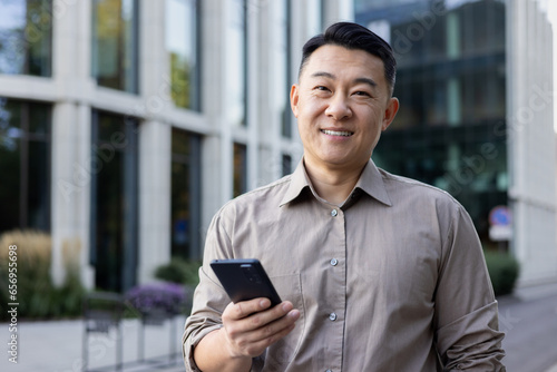 Portrait of Asian young man businessman, worker, manager standing outside office center and using mobile phone. Smiling at the camera. Close-up photo © Tetiana