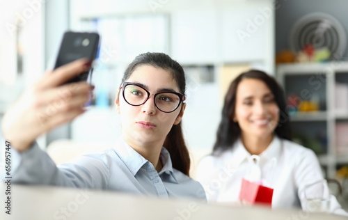 Young woman in glasses holds phone in front of face and smiles at colleagues in office. Video call or selfie at work