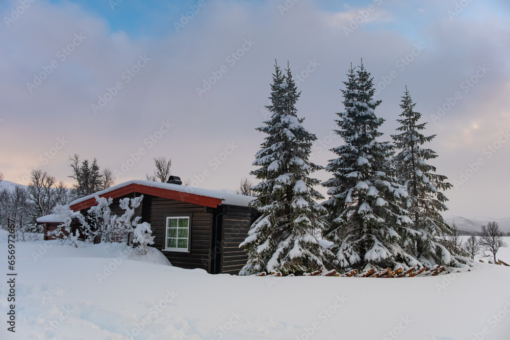 A small snow covered hunting lodge in the norwegian mountains with animal traces in the foreground and mountain summits in the background