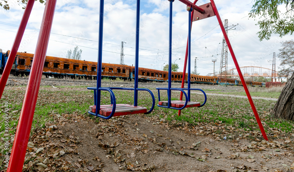 empty swings on an empty playground without people against the background of burnt trains in Ukraine