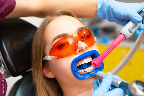 Patient in protective glasses from UV light with tubule for liquid suction. Dentist puts tubule in woman mouth to work on teeth and cavity cure