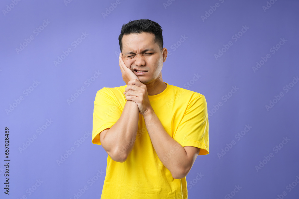 Portrait of young Asian man touch cheek. He has toothache and suffering from pain, wearing a yellow t-shirt isolated on a purple background. Portrait of young Asian man touch cheek. He has toothache and suffering from pain, wearing a yellow t-shirt isolated on a purple background.