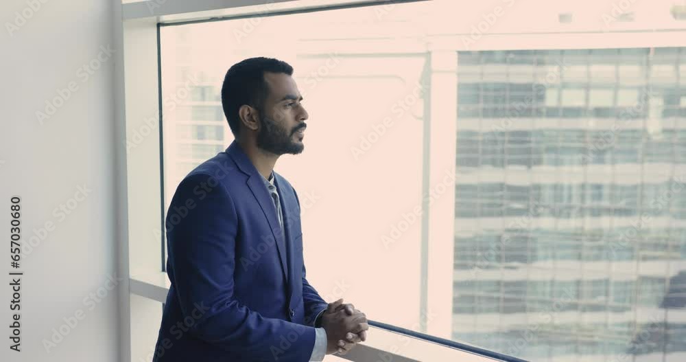 Young thoughtful Indian businessman in suit stand in modern skyscraper office, look out window, thinks of future, deep in thoughts about start up ideas, goals or project. Business vision, aspirations Young thoughtful Indian businessman in suit stand in modern skyscraper office, look out window, thinks of future, deep in thoughts about start up ideas, goals or project. Business vision, aspirations