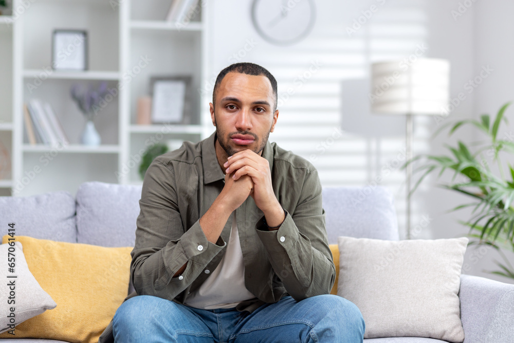 Portrait of a young hispanic man looking seriously at the camera, sitting at home on the couch with folded hands