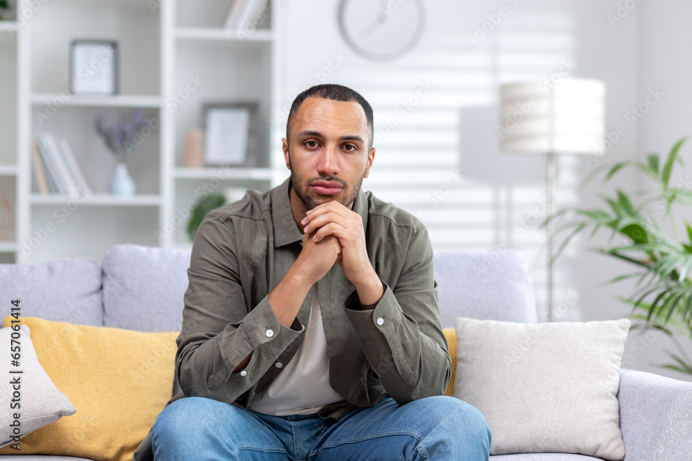 Portrait of a young hispanic man looking seriously at the camera, sitting at home on the couch with folded hands Portrait of a young hispanic man looking seriously at the camera, sitting at home on the couch with folded hands