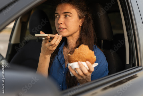 Close-up of woman using mobile phone, eating burger and talking on the speaker while driving car. Beautiful woman talking on in-car speakerphone