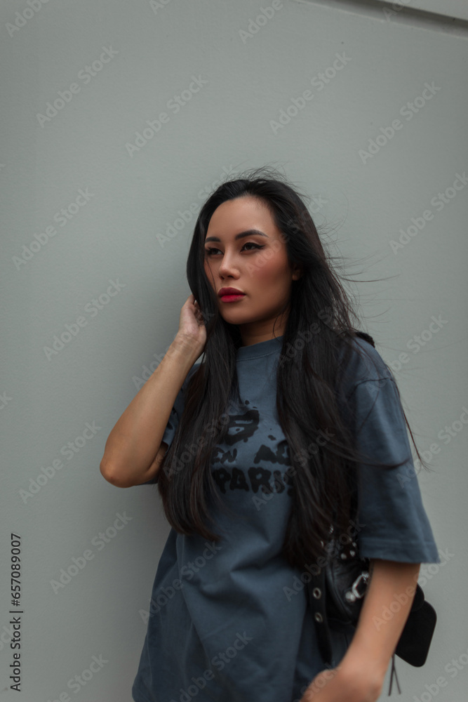 beautiful cool stylish Asian woman with red lips in a fashion urban outfit with a leather bag walks on a windy day in the city near a wall beautiful cool stylish Asian woman with red lips in a fashion urban outfit with a leather bag walks on a windy day in the city near a wall