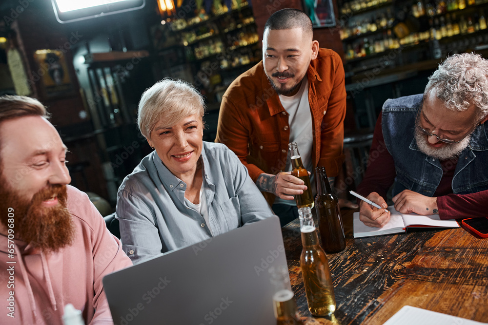 bearded businessman writing in notebook near multiethnic team talking near beer and laptop in pub bearded businessman writing in notebook near multiethnic team talking near beer and laptop in pub