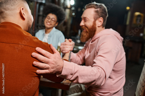 pleased bearded man shaking hands with colleague in pub near blurred african american woman