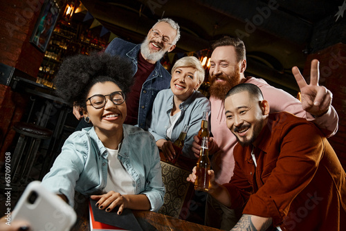 cheerful african american woman taking photo with multiethnic colleagues holding beer bottles in pub