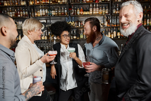 bearded tattooed man talking to smiling african american woman near workmates with drinks in bar