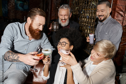 bearded tattooed man clinking glasses with multiethnic women near smiling workmates in cocktail bar