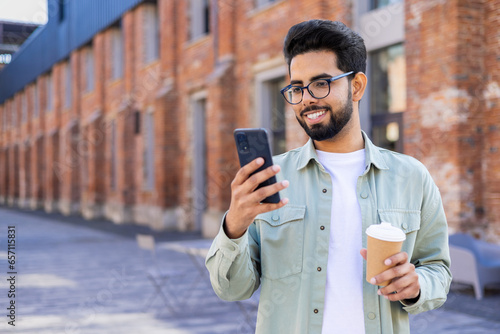 A young student uses the phone, the man holds a smartphone and a cup of hot drink in his hands, walks around the city, smiles, reads messages and watches online videos.