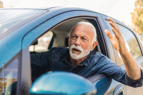 An irritated senior man driving a vehicle is expressing his road rage. Angry white hair man driving a vehicle. Front view of annoyed male entrepreneur screaming while driving a car during the day.
