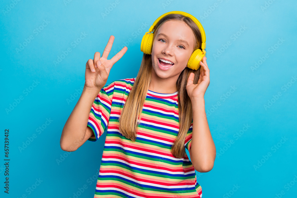 Portrait of good mood positive teen girl wear colorful t-shirt in headphones showing v-sign symbol isolated on blue color background Portrait of good mood positive teen girl wear colorful t-shirt in headphones showing v-sign symbol isolated on blue color background
