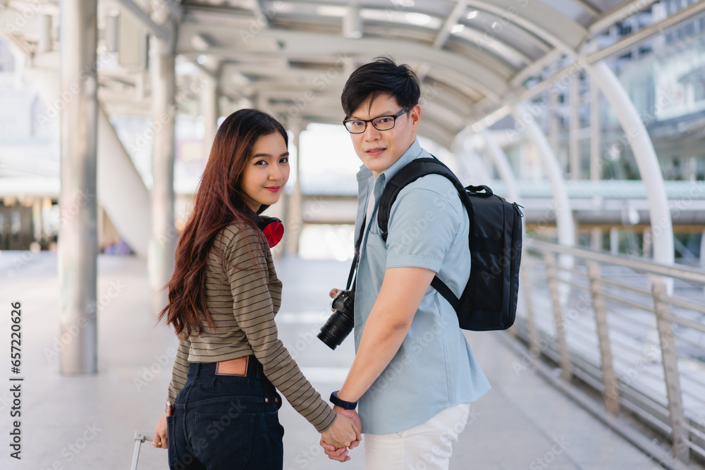 Asian tourist couple standing with luggage before going on summer vacation or honeymoon trip in airport or bus station or train station. A couple is about to walk hand in hand, a romantic atmosphere.