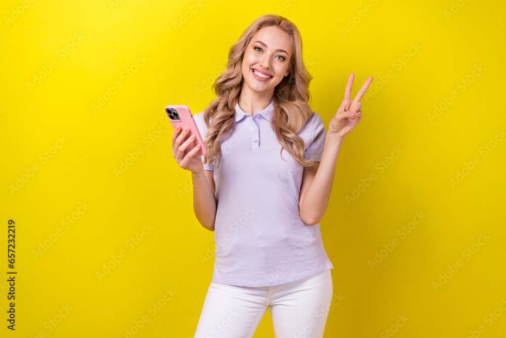 Photo of positive gorgeous girl toothy smile hold smart phone demonstrate v-sign isolated on yellow color background Photo of positive gorgeous girl toothy smile hold smart phone demonstrate v-sign isolated on yellow color background