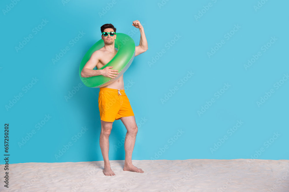Full size photo of attractive young man hold float circle raise fist excited wear trendy yellow shorts isolated on blue color background Full size photo of attractive young man hold float circle raise fist excited wear trendy yellow shorts isolated on blue color background